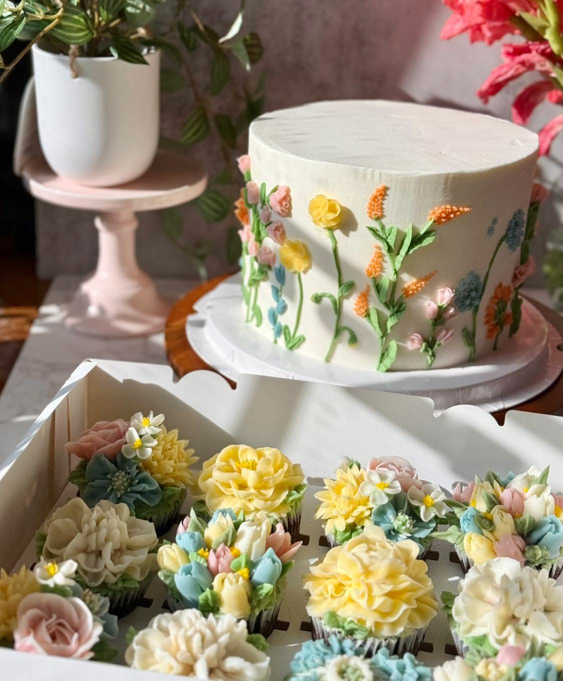 White buttercream cake hand-piped with a meadow of wildflowers, beside a box of floral cupcakes